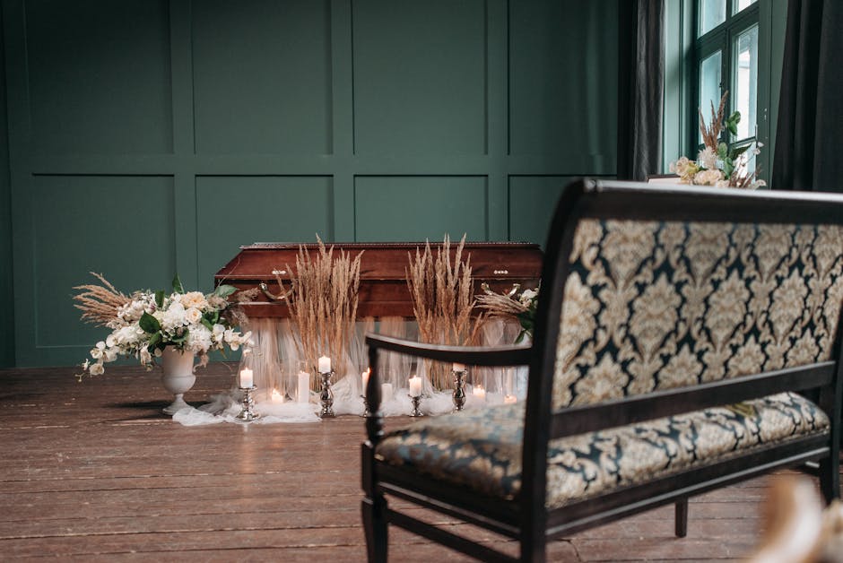 An elegant floral display arranged around a wooden coffin, featuring dried beige pampas grass and soft white blossoms arranged in white ceramic vases on either side. The arrangement is accented with s
