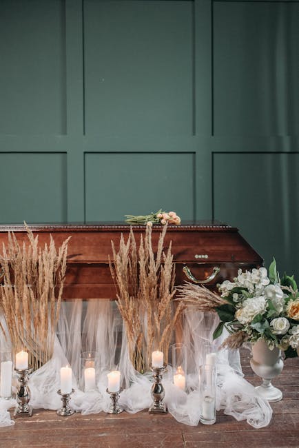 A floral tribute featuring a bouquet of light pink roses and white flowers, resting on a polished wooden casket with a dark finish. The display is decorated with tall, golden-hued pampas grass and ill