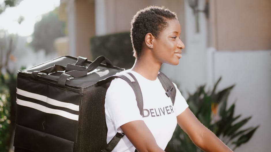 A smiling woman with short, natural hair is outdoors during daytime, wearing a white T-shirt with the word 'DELIVER' printed on it and a grey backpack by her shoulders. She appears to be a delivery pe