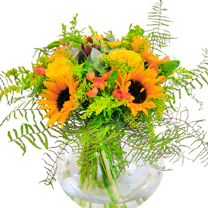 First impressions of this exuberant bouquet are a rush of sunlight and cheerful color, as if the vibrant spirit of Crystal Palace itself has been distilled into petals and greenery. Tall, radiant sunflowers and lush yellow roses bloom side by side, their layered petals creating a feeling of generosity and warmth. Threaded between are deep emerald ferns and light, airy foliage, tumbling outward like the leafy avenues around Anerley Hill. Crimson clusters of hypericum berries gleam within the bouquet, offering bold contrast, much like the surprising pops of colour from local street art. Delicate stems of goldenrod mingle with round blue-green eucalyptus leaves, adding subtle scents of the season. The clear, fishbowl-like vase showcases the stems beneath-clean, structured, and always fresh, as expected from Crystal Palace's dedicated florists. Studying it closely, you'd notice velvet-soft sunflower faces, the intricate swirl of rose petals, and the crisp snap of new greenery. This bouquet's gentle fragrance and vibrant look evoke the open fields at the edge of the park, making it an ideal gift for brightening birthdays, marking anniversaries, or simply spreading happiness from Upper Norwood to Gipsy Hill.